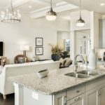 Custom designed kitchen island with marble counter top, double sink, metal hanging light fixtures and living room with white couch and wooden tv stand.