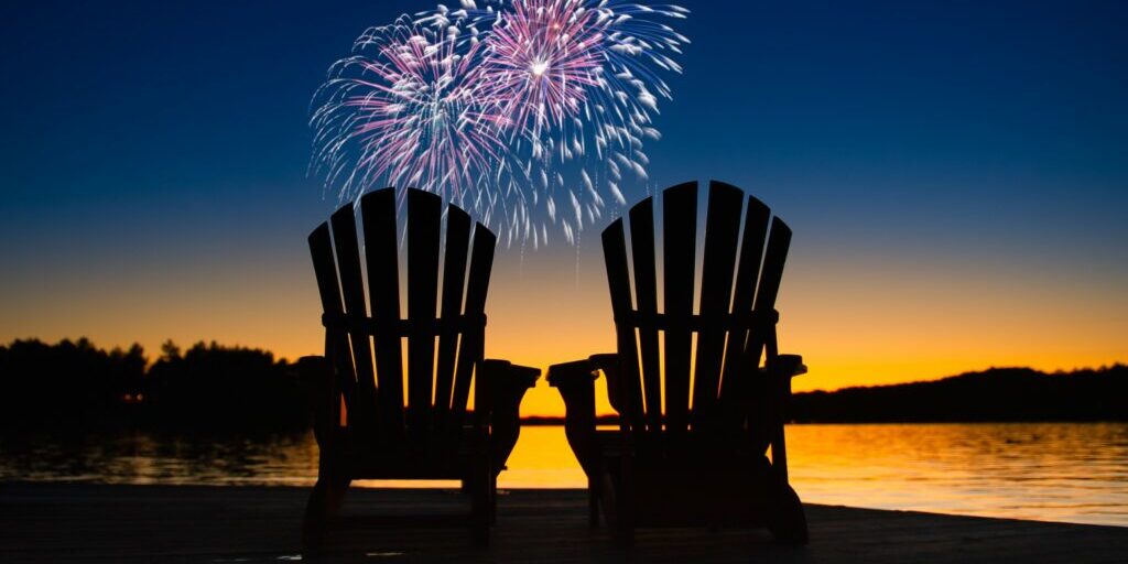 Canada day fireworks on a lake in Muskoka, Ontario Canada. On the wooden dock two Adirondack chairs are facing the sunset orange hues